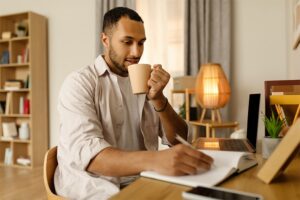 Man writing in notebook while holding a mug