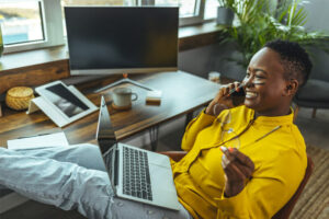 African American lady with a laptop sitting at her desk while talking over a smartphone