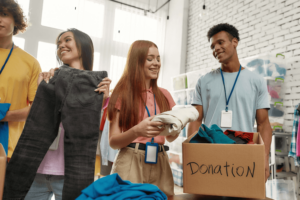 Happy young volunteers sorting, packing clothes in cardboard boxes, Diverse team working together on donation project indoors.