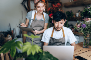 Young Asian couple, owners of small business flower shop, using laptop and working together in the shop against flowers and plants.