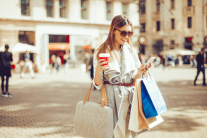 Cheerful woman is shopping in the city. She is holding a coffee to go and using her smart phone.