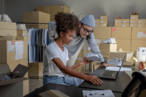 Mixed race couple of Asian man and African-American woman online seller confirming orders from customer on laptop. E-commerce business owner looking at laptop in store warehouse.