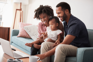 Family With Baby Daughter Sitting On Sofa At Home Looking At Laptop Computer.