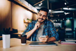 Smiling hipster guy reading new message in social networks confirming new friend on smartphone, positive young man satisfied with banking app on mobile phone checking balance while learning.