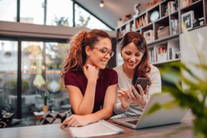 Two smiling woman looking at smart phone while sitting a desk with laptop, in modern house, portrait.