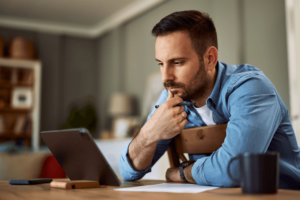 A deeply focused male freelancer looking at a tablet with a hand on his chin and sitting backwards in a chair.