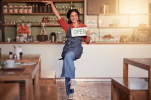 A happy female business owner in her cafe. She stands on one foot in a happy dance move, holding a sign that says "Open."