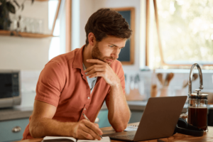 Young caucasian businessman writing in a notebook and working on a laptop at home alone.