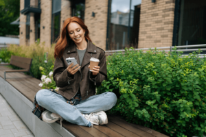 Portrait of joyful redhead female in jacket sitting on urban bench, enjoying cup of coffee and using mobile phone, taking break from day to relax.